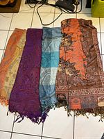 Four scarves laid out side by side showing paisley and floral patterns in orange, purple, blue, and multicolor hues with fringe.