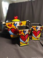 Front view of the teapot and two matching mugs on black background showing colorful Britto design and teapot lid with red heart knob