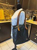 Front side view of black leather tote bag with tan handles displayed on a mannequin torso in kitchen setting