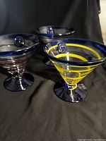 Three handblown Kosta Boda Royal Caribbean Swirl cocktail glasses with cobalt blue rims and bases, two with burgundy swirls and one with yellow swirl, on black background.