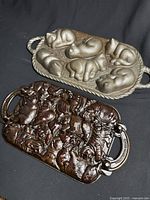 Two John Wright cast iron baking molds displayed together on black cloth, showing the animal cookie mold with multiple animal figures and the cat-shaped muffin pan with dark seasoned cast iron patina.