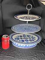 Front view showing three porcelain plates stacked with intricate blue floral and geometric patterns and a silver-tone metal handle on top. Coke can shown for size reference.