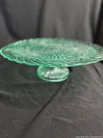 Side view of green pressed glass cake stand on a black background showing scalloped edge and pedestal base
