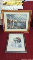 Two framed nature prints by Al Poolman displayed on a red surface with textiles above them. The larger wooden frame print features a scene of birch trees by a lake.