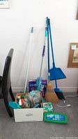Full lot view showing brooms, mops, box of supplies, and Swiffer refills arranged against a wall.