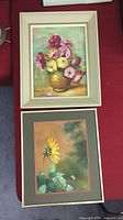 Photo of two framed floral paintings arranged on a red surface. One painting shows pink and yellow flowers in a vase; the other is a sunflower with green leaves and a butterfly.