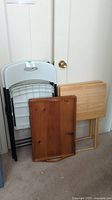 View of two plastic folding chairs, two wooden folding tables, and a wooden serving tray stacked upright against a wall