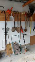 Wide view of gardening tools hanging on wall including shovel, hand tools, dustpan, and electric weed whacker at bottom right