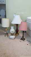 Four table lamps positioned on carpet showing bases and shades with a mattress to the side and bathroom in background