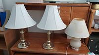 Three table lamps displayed together. Two lamps have brass bases with adjustable arms and clean white shades, the third lamp has a rounded white ceramic base and a white shade with a stain.