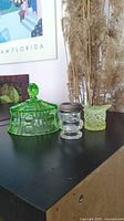 Three glass items on black surface: a green uranium glass hat figurine, a clear glass dish with silver rim, and a green uranium glass candy dish lid.