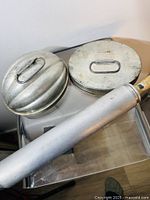 Photo showing two KREAMER tin pudding molds and antique aluminium rolling pin on clear plastic stand against a white wall.