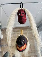 Two miniature Indigenous Algonquin Iroquois carved false face masks shown hanging, highlighting colors and attached long white hair.
