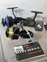 Two vintage fishing spinning reels displayed together, one Garcia with blue spool and one black Mitchell with fishing line and gold label. Mitchell manual in foreground.