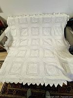 Full view of vintage white crocheted knitted bedspread blanket spread over couch showing square crochet motifs and scalloped edges.