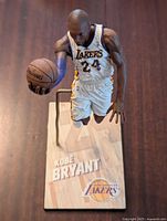 Front view of Kobe Bryant figure in mid-dunk pose holding basketball, wearing white Lakers uniform number 24, mounted on a stand with base showing Kobe Bryant name and Lakers logo.
