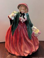 Front and side view of the Lady Charmian figurine showing red dress, green cape, bonnet, and flowers in hand and basket
