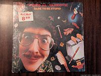 Front cover of Weird Al Yankovic Dare to Be Stupid vinyl record showing artist's face and colorful objects floating around.