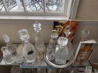 Overall view of six cut glass decanters with stoppers, glass pitchers and silver-tone tray on glass shelf