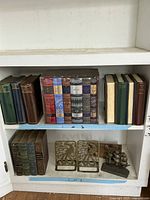 Shelf with hardcover classic literature books arranged neatly in two rows. Bottom row displays decorative metal bird-themed bookends and a sailing ship tissue box cover.