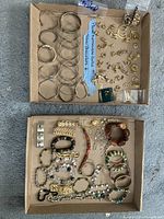 Two open cardboard boxes with various bracelets and gold plated earrings spread inside. Bracelets are silver tone bangles and different style chain and beaded bracelets. Earrings include gold plated studs, hoops, and decorative shaped pieces.