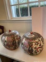 Two round Chinese ginger jars with lids on white table near window, decorated in detailed Rose Medallion style with birds, flowers, Asian scenes.