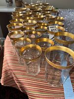 Angle view of clear glassware with wide gold rims showing multiple cups arranged in rows on a striped cloth.