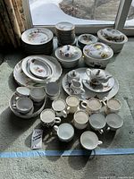 Full set of dinnerware including plates, cups, saucers, covered serving bowls, teapot, displayed on carpeted floor with natural light.