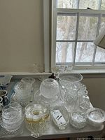 Top view showing full collection of assorted clear cut glass dinnerware including bowls, plates, vases, and serving pieces arranged on a table near a window.