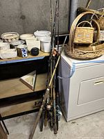 Eight fishing rods standing against a dryer and shelf with baskets and planters on top.