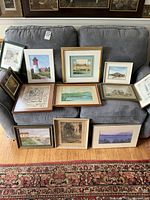 Wide group shot of 13 framed prints on a couch and floor, showing mix of coastal scenes and etching.