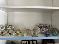 Full shelf view showing all 11 glass paperweights in various designs and shapes including cats, flowers, and fish.