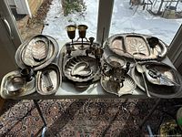 Photo showing entire lot displayed on a table, includes multiple silver-plated trays, candle sticks, baskets, salt and pepper shakers, serving utensils, and bud vase.