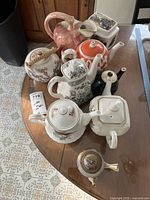 Top-down photo showing a group of seven teapots and a rectangular tea bag jar on a glass-top table. Teapots show various styles, including floral, oriental, and animal decorations.