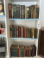 Three rows of vintage hardcover books on white shelves, showcasing a variety of classic titles and editions with mostly green and brown covers.