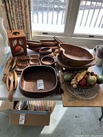 Wide view showing an assortment of wooden bowls, utensils, a cutting board, artificial fruit bowl and wooden candle holder on a table.