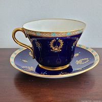 Side angle of teacup and saucer showing cobalt blue surface, gold wreath designs and raised turquoise dots, cup handle with thick gold detail