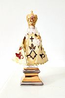 Front view of the porcelain figurine of Infant Jesus of Prague with fabric embroidered vestment and gold crown, standing on metallic pedestal base.