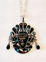 Close-up photo of aztec style pendant with carved black mask face and colorful inlaid gemstones on headdress with dangle accents