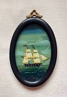 Front view of the reverse glass painting of a ship in an oval black wood frame with a brass hanging hook.