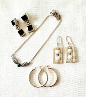 Top view of assorted sterling silver jewelry, including square onyx earrings, a twisted silver necklace with green stone, rectangular pearl dangle earrings, and textured hoop earrings.