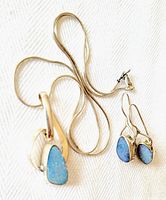 Photo showing the necklace with pendant and the earrings arranged on a white textured surface, displaying the blue opalescent stones and the silver settings.