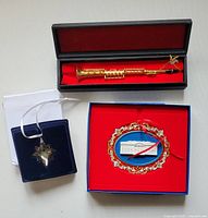 Photo showing all three ornaments together: gold metal clarinet in black box, Swarovski crystal snowflake in blue velvet box, and White House ornament in red box.