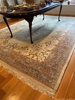 Photo showing the corner of the area rug under a dark wooden table, revealing the floral vine design in beige, dusty blue and muted red tones along with the fringed edge.