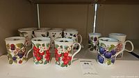 Shelf display of 14 porcelain mugs with fruit designs including cherries, strawberries, plums, blackberries.