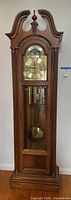 Full front view of the wooden grandfather clock showing the swan-neck pediment, glass door, weights and pendulum.