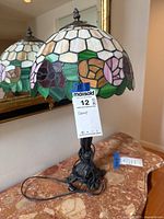 Photo showing the entire stained glass floral table lamp with its metal figural base on a marble surface with cord wrapped around the base.