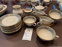 Overview of stacked saucers at left, teacups, sugar bowl with lid and creamer at right with lot tag