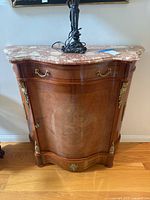 Front view of the serpentine front credenza showing drawer with brass handles, curved cabinet door, decorative brass mounts at corners, and reddish-pink marble top with veining. Black lamp base visible on top (lamp not included).