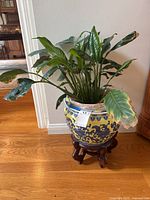 Full view of ceramic planter with live plant on wooden stand in room with hardwood floor and partial furniture in background.